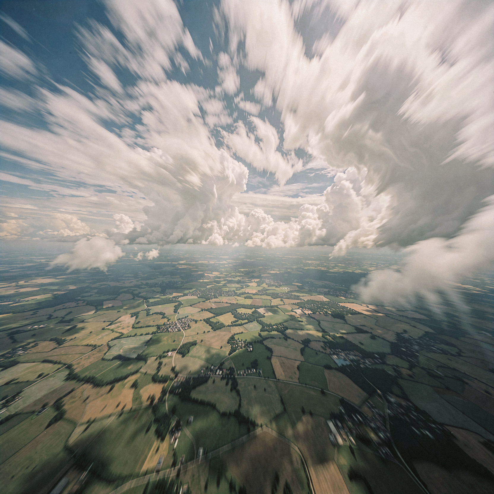 a realistic aerial photograph looking down from a great height, capturing the sensation of weightless flight above a vast patchwork of earth and cloud. The viewer is alone in the frame, suspended in a state of serene equilibrium where the ground below recedes into an abstract tapestry of greens and browns, while the sky above swirls with turbulent, energetic clouds that suggest wild freedom rather than danger. The lighting is bright and expansive, creating a sense of infinite space and quiet solitude without loneliness, blurring the line between dream logic and perceived reality as the horizon curves gently into the atmosphere. Shot on medium-format film with soft grain and gentle motion blur in the cloud formations to convey the feeling of drifting, natural unretouched skin texture visible on hands gripping the air, slight handheld framing imperfection suggesting a personal, intimate perspective.