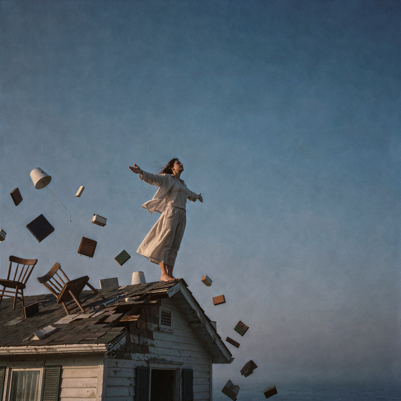 a realistic editorial photograph of a solitary woman standing on the edge of a crumbling suburban roof that dissolves into an endless, cloudless azure sky, her arms spread wide to embrace the overwhelming void. The house below is merely a small platform of weathered shingles and siding, surrounded by floating debris of domestic life—chairs, lamps, books—drifting slowly upward into the infinite atmosphere, symbolizing the shedding of confinement for boundless freedom. She wears light linen clothing that flutters in a steady thermal updraft, her expression one of serene release and quiet awe as she gazes toward the horizon where the sky deepens to indigo. The composition emphasizes vast negative space above and around her, reinforcing the feeling of expansion and liberation from earthly constraints. Soft natural daylight illuminates the scene from camera-left, casting gentle shadows and highlighting the texture of the fabric and skin. shot on medium-format Portra 160, soft grain and gentle highlight rolloff, slight handheld framing imperfection, atmospheric haze in the distance