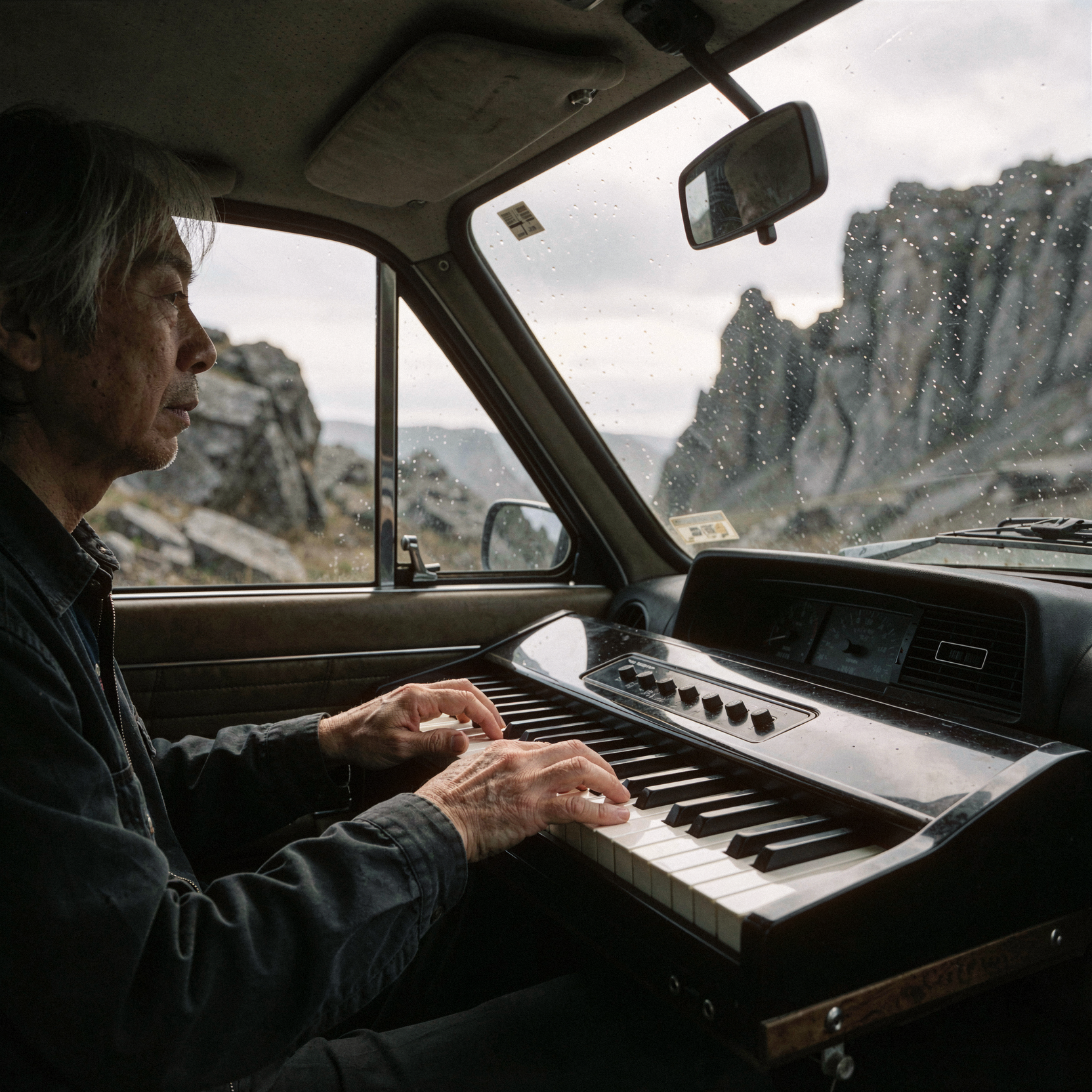 a realistic documentary photograph of Ryuichi Sakamoto seated inside a vintage car, playing a compact portable electric piano mounted on the dashboard. his fingers press the keys with gentle intensity as he gazes forward through the windshield toward a vast, rugged landscape of jagged grey rocks and steep cliffs under an overcast sky. the interior of the car is dimly lit by ambient daylight filtering through the windows, casting soft shadows across his face and the instrument's glossy surface. dust motes float in the air between him and the glass, blurring slightly in the shallow depth of field. shot on 35mm Kodak Portra 400 with fine visible film grain, slight handheld framing imperfection, natural unretouched skin texture with subtle tonal variation, realistic highlights on the forehead, smooth continuous tonal gradients in the sky, clean continuous tonal gradients in the rock formations, soft diffuse edges on the distant landscape.